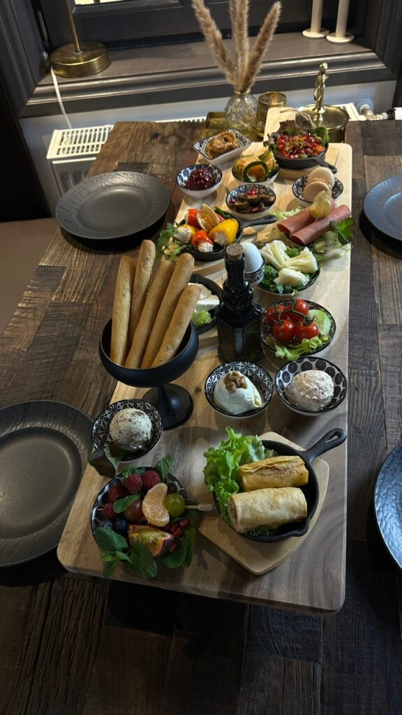 Auf einem rustikalen Holztisch steht eine lange Holzplatte mit verschiedenen köstlichen Häppchen wie Baguette, Käse, Obst, Dips und Gemüse, arrangiert mit schwarzem Geschirr und eleganten Schalen in dunklen Farben