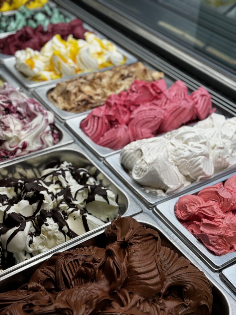 Mehrere Behälter mit verschieden farbigem cremigem Eis sind in einer Vitrine ausgestellt
