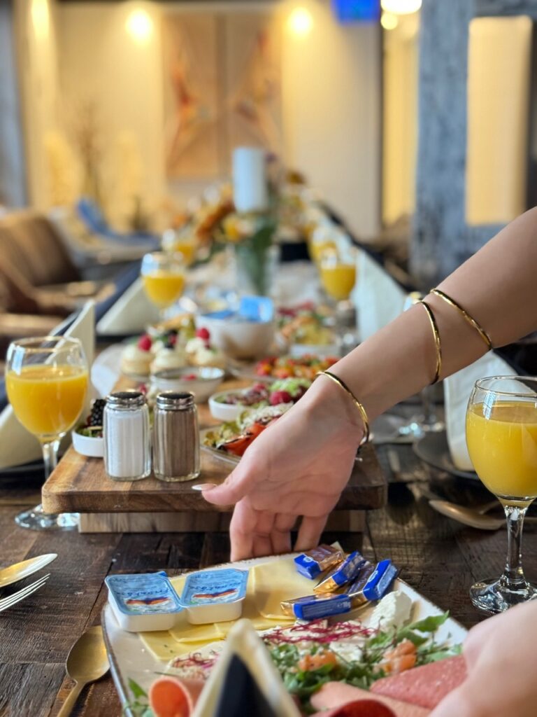 Eine Frau mit goldenen Armreifen reicht am elegant gedeckten Frühstückstisch Käse weiter